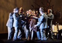 Nine men in military uniforms holding glasses restrain a woman in a floral dress who recoils with a look of terror.